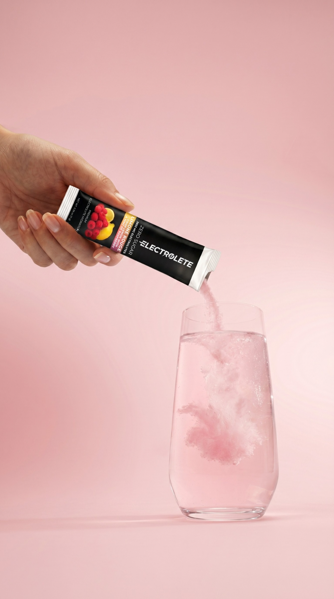 Hand holding an ELECTROLETE drink mix packet over a glass of pink liquid on a pink background